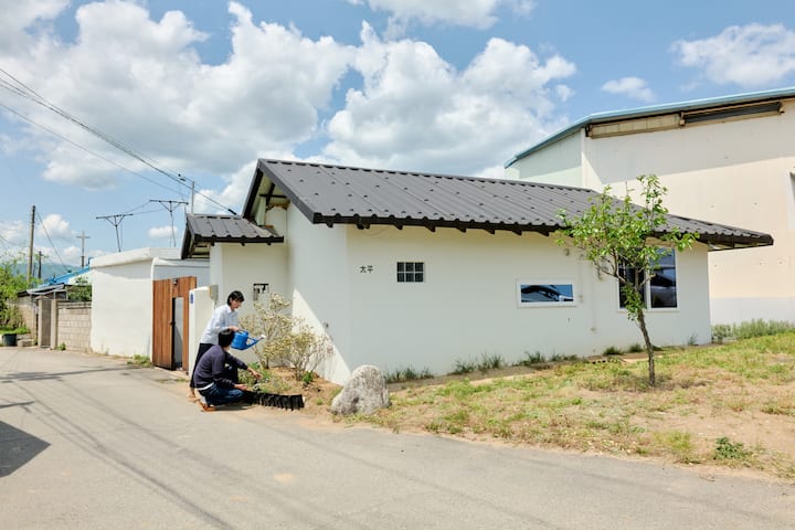 Goseong host tending the garden outside a small white house under a bright blue sky
