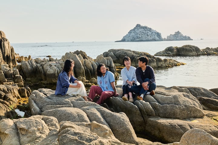 Four friends sitting on seaside rocks in Goseong, smiling and talking by the calm water