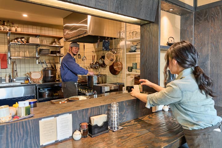 Goseong host cooking in a cozy open kitchen, smiling as a guest takes a photo from the counter
