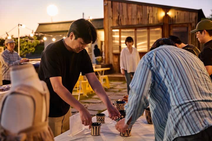 Host in Goseong arranging striped ceramic cups on an outdoor table at sunset gathering