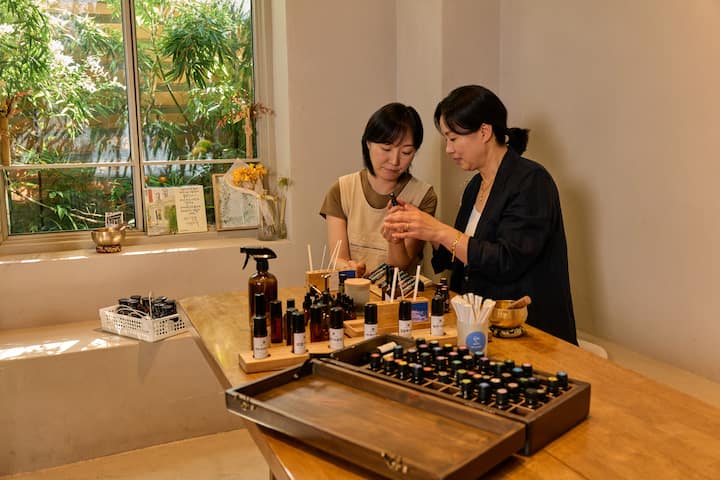 Host in Suncheon guiding guest through a table of essential oils and aromatherapy bottles in a bright studio.