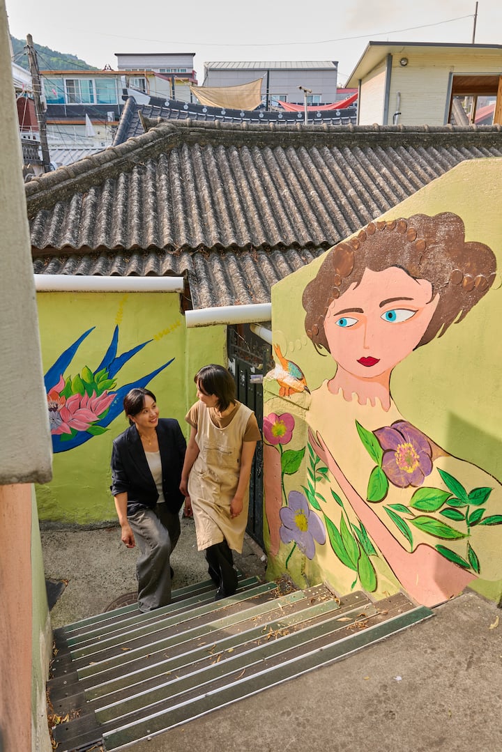 Two Suncheon hosts chatting on colorful mural-lined stairs in a hillside neighborhood
