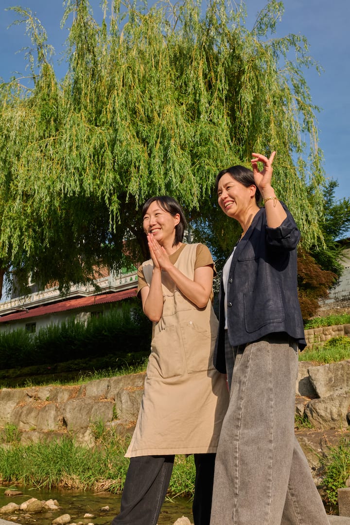 Two cheerful Suncheon hosts share a moment outdoors beside a creek and greenery on a sunny day.
