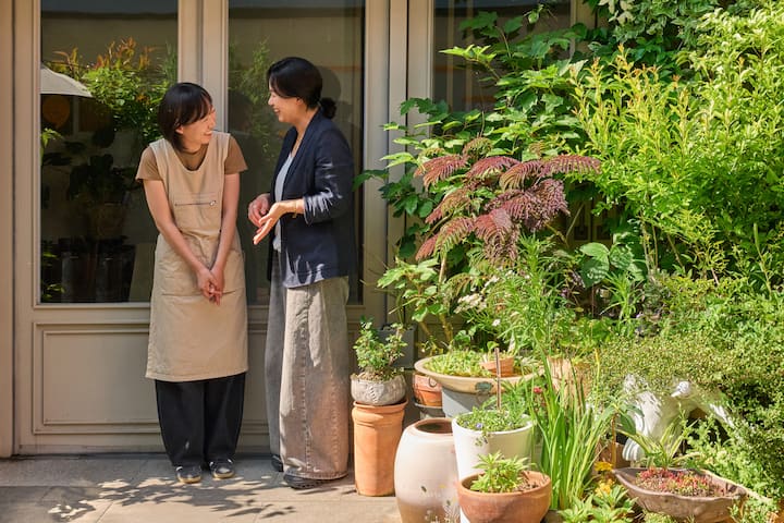 Suncheon host chatting with a guest beside lush potted plants outside a home entrance