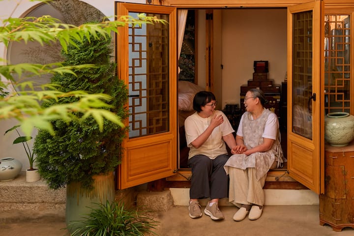 Seongbuk-dong host sharing conversation with a woman outside a serene courtyard room, framed by plants and wooden doors