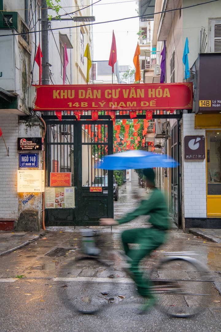 Cyclist with blue umbrella rides past the 14B Lý Nam Đế residential gate in rainy Hanoi