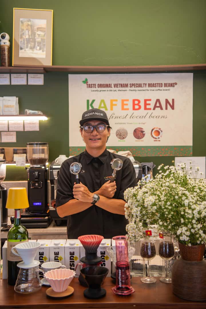 A man smiles behind a Kafebean counter, holding espresso portafilters amid local coffee gear.