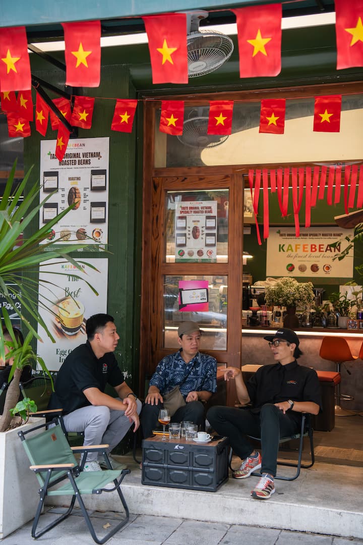 Vietnamese host Khoi chats with two friends outside his flag‑decorated café in Vietnam.