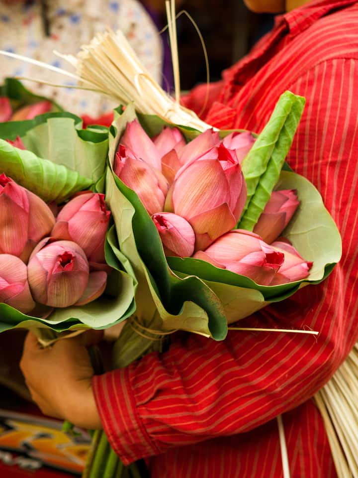 Person in a red shirt holding large bundles of pink lotus flowers wrapped in green leaves in Vietnam