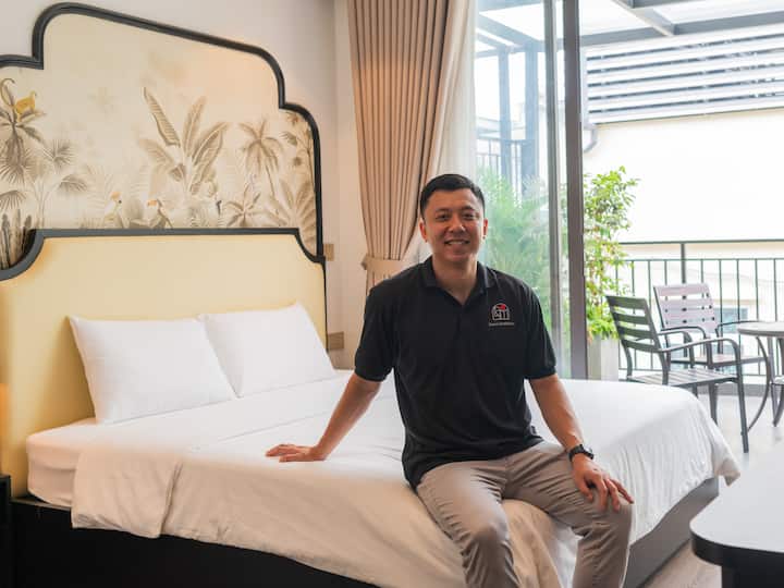 Khoi sits smiling on a neatly made bed in his bright Airbnb room with balcony patio.