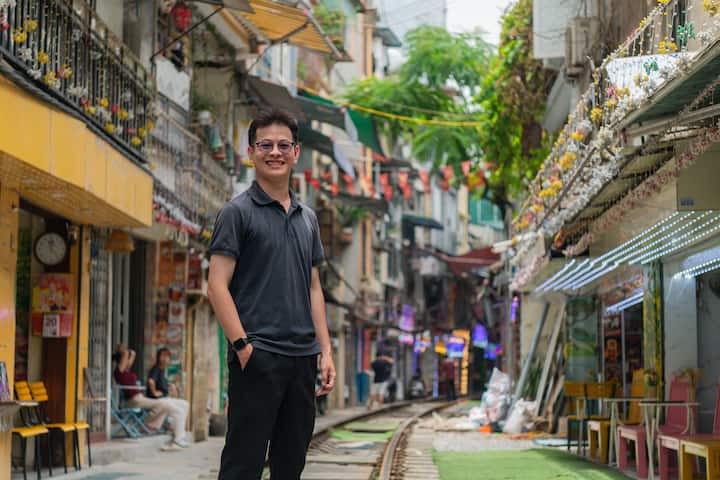 Quang standing beside Hanoi train street, smiling in front of cafés and homes lining the historic tracks