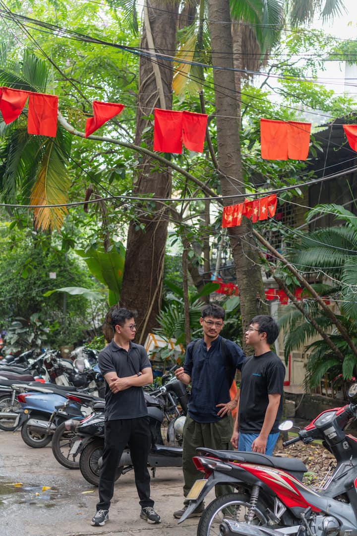 Vietnamese hosts talking by motorbikes under red festival flags in a lush neighborhood courtyard.