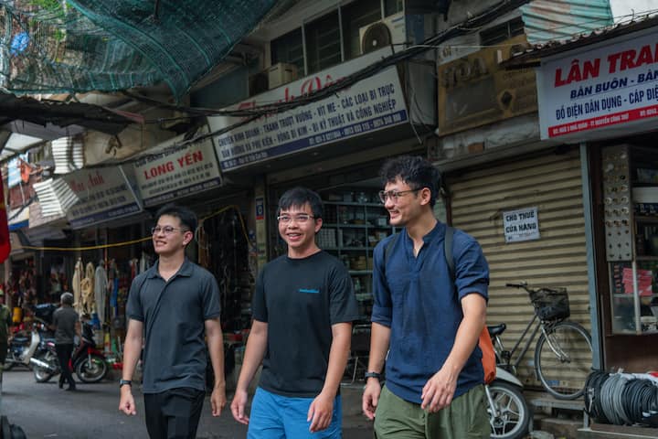 Quang walks with two guests through Chợ Giời market street in Hanoi, passing small local shops.