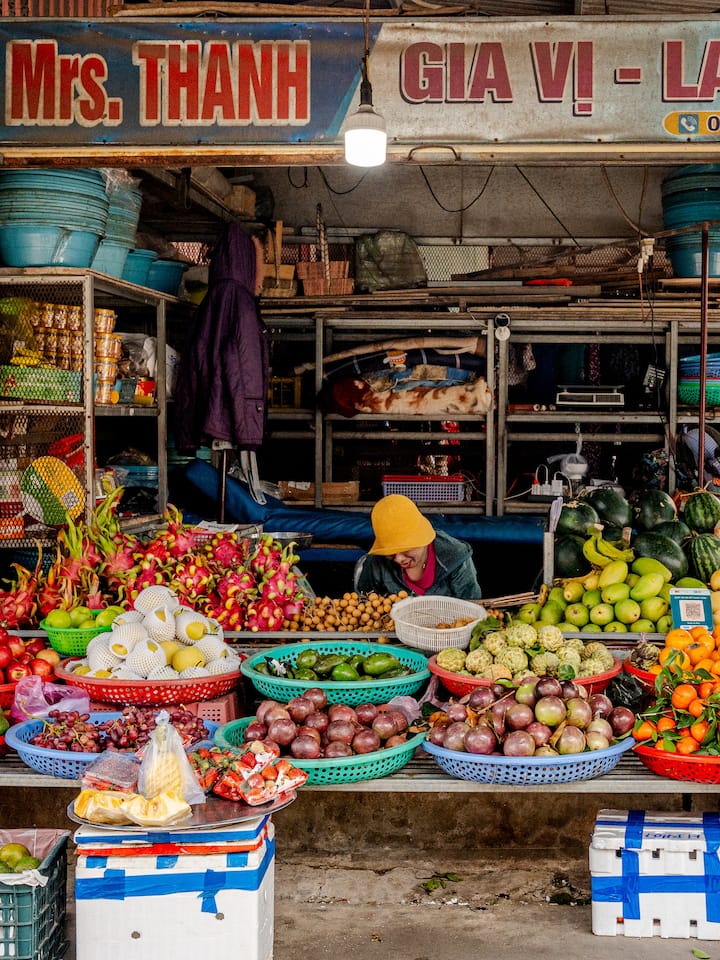 Vietnamese market stall overflowing with colorful fruit as a local vendor in a yellow hat arranges produce.