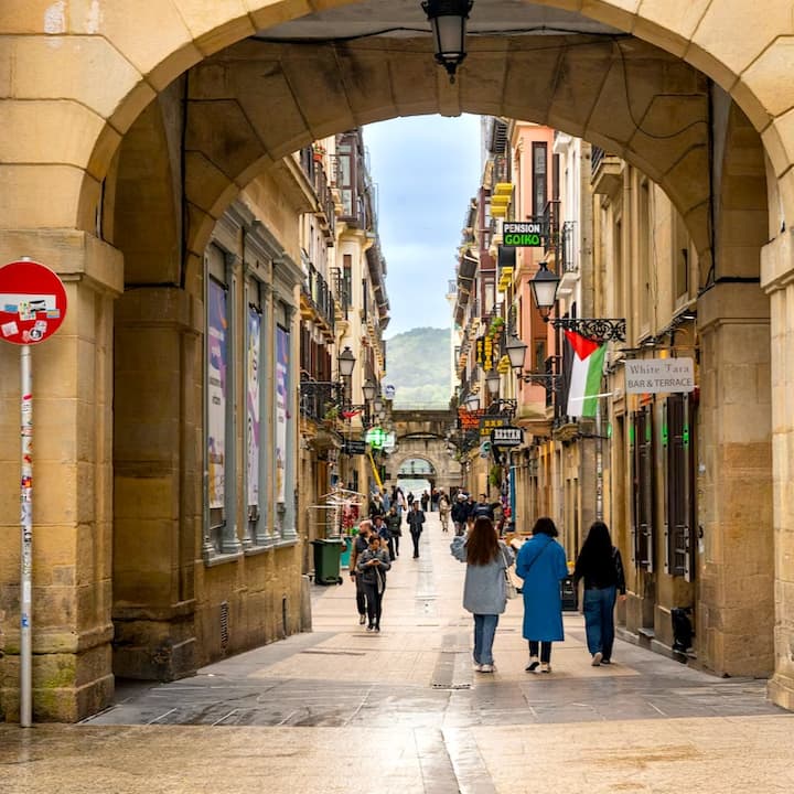 Personas paseando por un callejón histórico de una localidad europea enmarcado por un arco de piedra, con cafeterías, letreros y colinas a lo lejos.