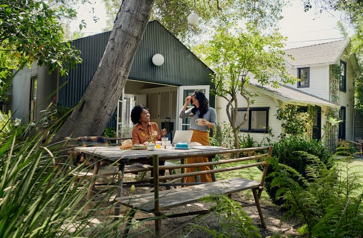 Two guests share breakfast at an outdoor table in a leafy garden beside a quiet Airbnb home.