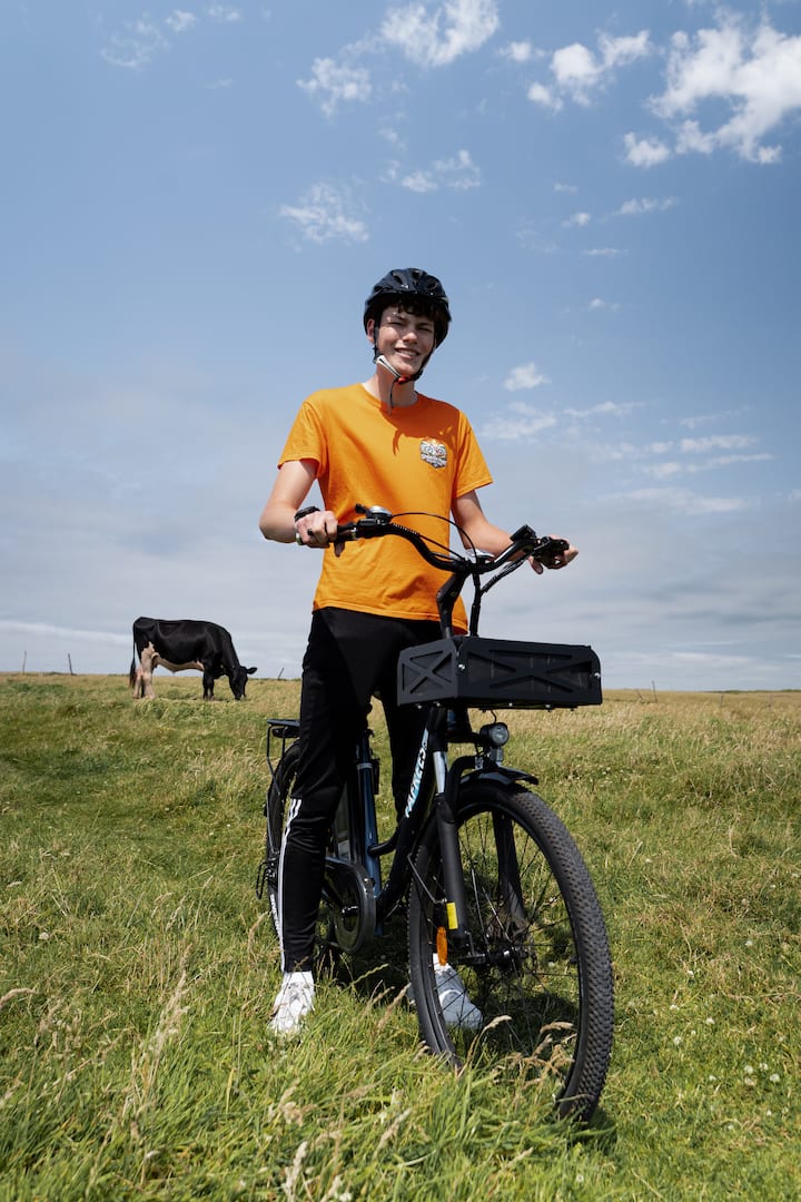 Smiling cyclist on a grassy rural trail with cows grazing under a bright blue sky