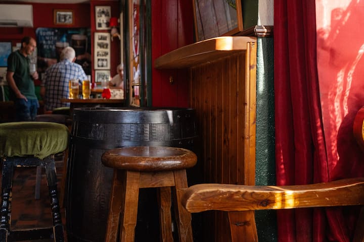 Cozy rural pub interior with wooden stools, barrel table, and locals chatting over pints in warm afternoon light