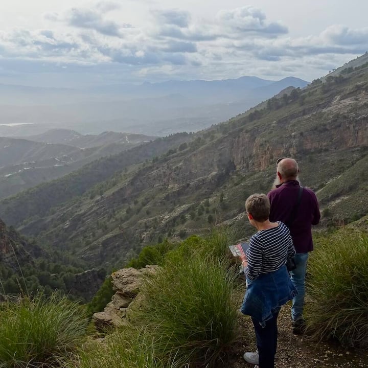 Una pareja hace una pausa en un estrecho sendero en la ladera de una colina para admirar el amplio paisaje montañoso y brumoso que se extiende a sus pies.