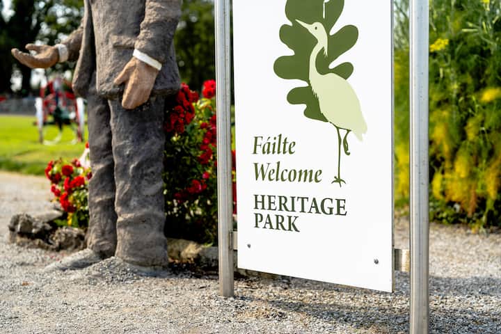 Sign reading “Fáilte Welcome Heritage Park” beside a rural path with flowers and greenery.