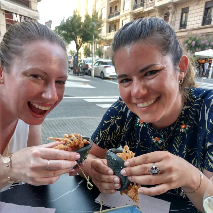 Dos amigas ríen juntas en la terraza de una cafetería durante una experiencia gastronómica al aire libre en Airbnb. Tienen unos tacos crujientes en las manos.