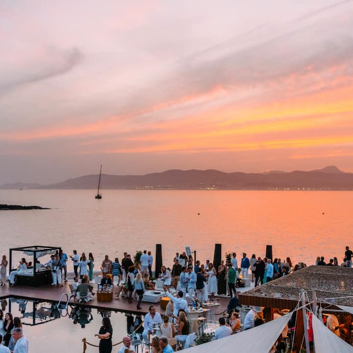 Varios viajeros están reunidos en una plataforma frente al mar al atardecer y charlan bajo un cielo naranja resplandeciente.