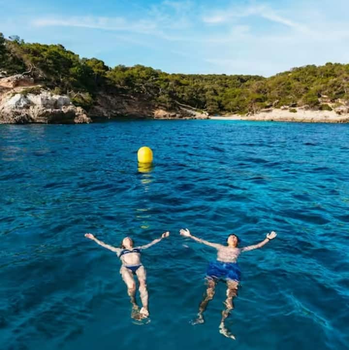 Unos viajeros están flotando en una bahía turquesa con los brazos extendidos. A lo lejos hay acantilados boscosos y una playa de arena.