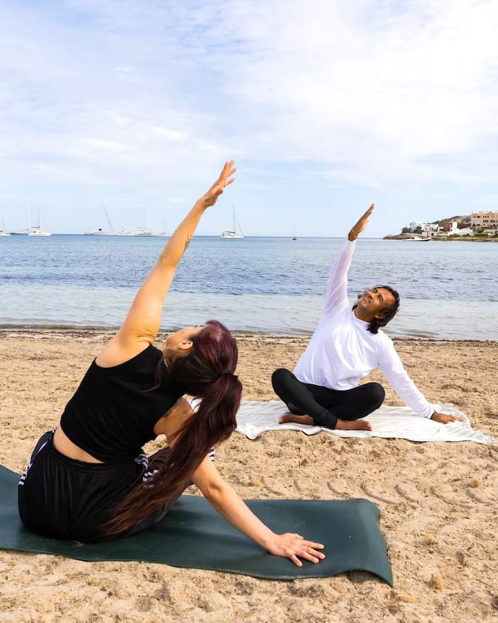 Dos personas practican yoga en una playa de arena en Ibiza. A sus espaldas hay veleros en el mar en calma.