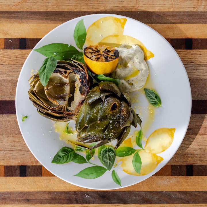 Chef-prepared grilled artichokes with charred lemon, sauce, and fresh basil plated on a wooden board