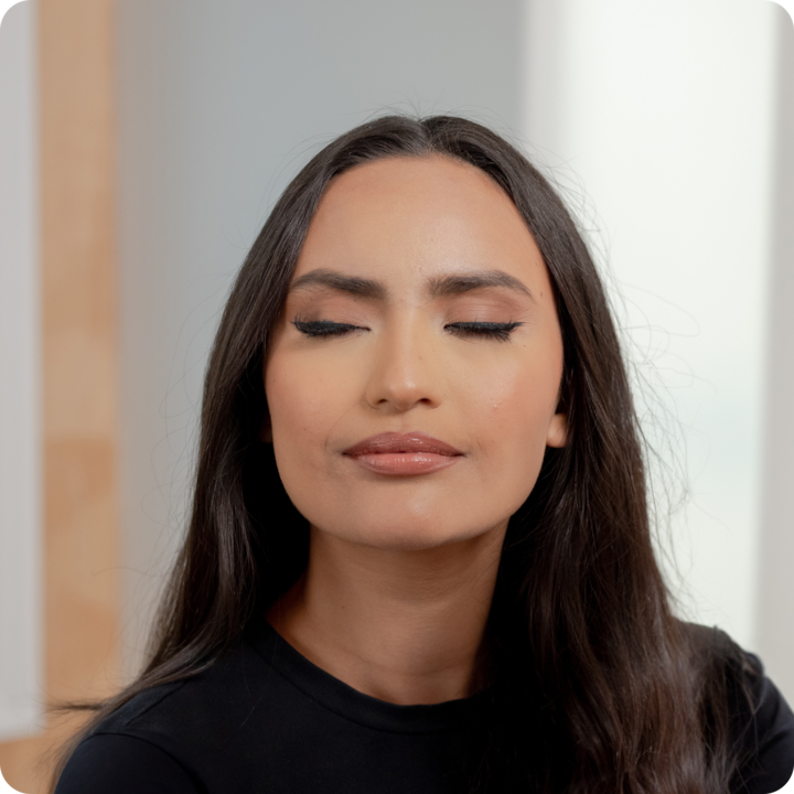 Young woman with long dark hair sits indoors with her eyes closed and a relaxed, content expression