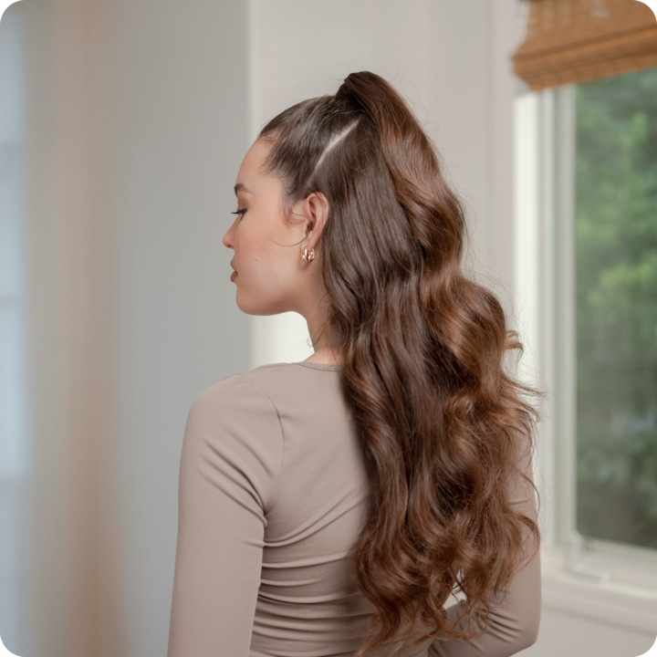 Stylist showcases a guest’s long, wavy ponytail hairstyle inside a bright home near a window.