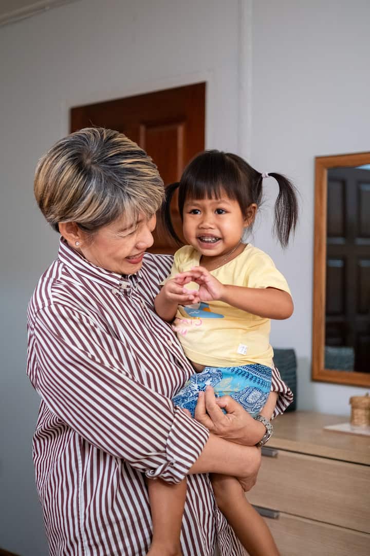 Smiling Thai host holding a young child in her home, sharing a warm moment together.