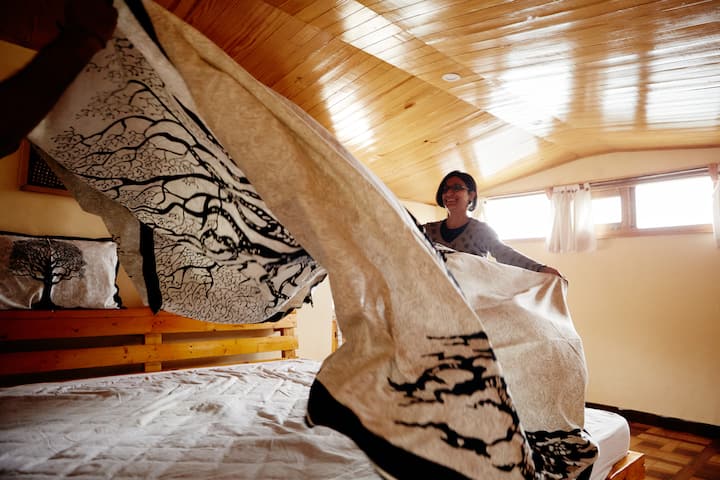 Host in a wood‑ceiling room spreads a tree‑patterned quilt over a guest bed, capturing the soul of India.