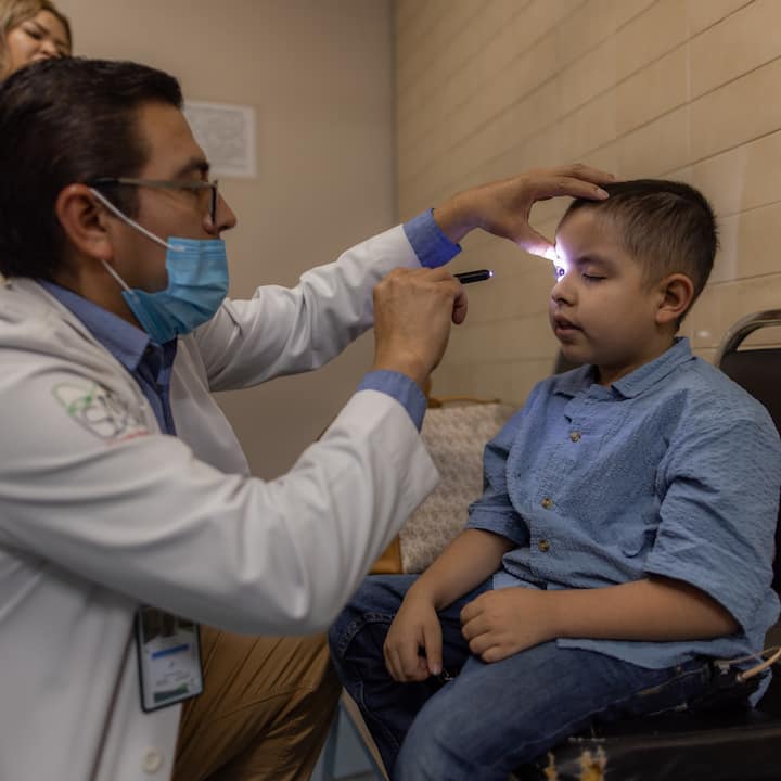 Un médico con bata blanca y cubrebocas examina con una lamparita el ojo de un niño, en una habitación con iluminación cálida.