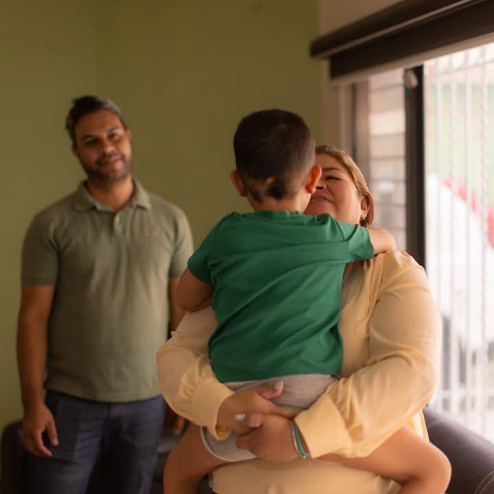 Un padre está de pie junto a una madre que carga con cariño a su hijo pequeño cerca de una ventana en el alojamiento que obtuvieron a través de Airbnb.org.