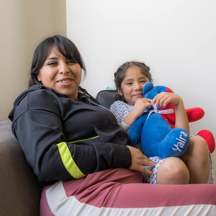 Una madre está sentada junto a su hija pequeña, quien lleva puesta una bata de hospital y abraza un peluche azul y rojo.