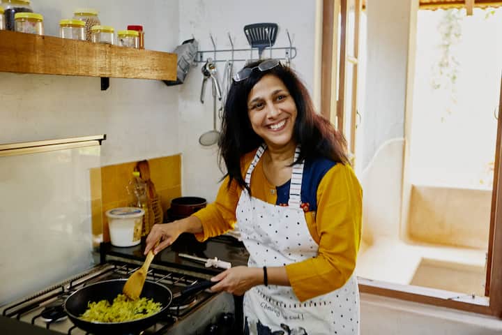 Host in a bright Indian kitchen smiling while cooking vegetables on the stove