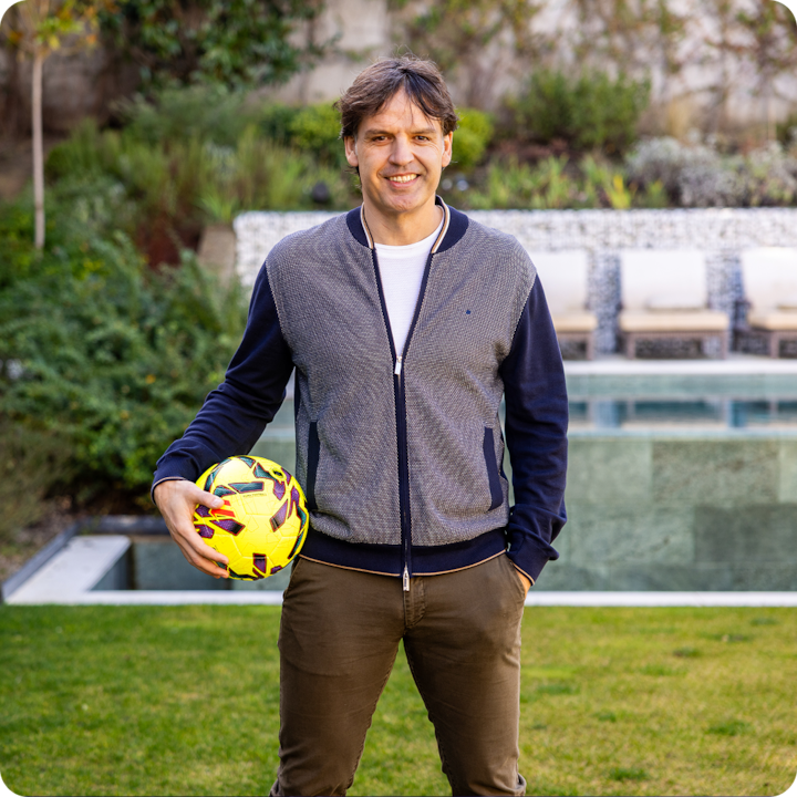 The footballer Fernando Morientes is smiling, holding a yellow football, standing outdoors in a landscaped garden by a swimming pool. He’s wearing a dark cardigan over a white shirt and brown trousers.