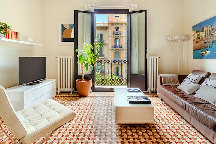 A bright living room with a patterned tile floor, brown leather sofa with pillows, white tufted lounge chair, white coffee table, TV on a white console, potted plant, and French doors opening to a Juliet balcony overlooking a European street with balconied buildings.