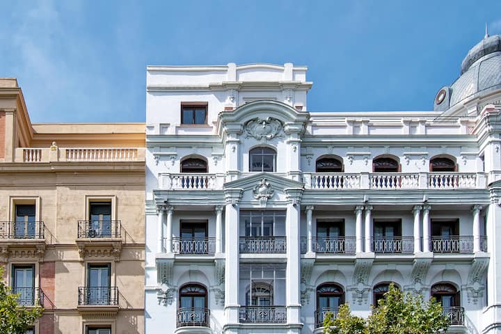 Facade of an ornate white classical building with balconies and decorative stone carvings, next to a tan-colored building, under a clear blue sky.