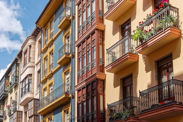 A row of colorful apartment buildings with wrought-iron balconies, wooden shutters, and balconies decorated with red flowers, viewed from below against a blue sky.