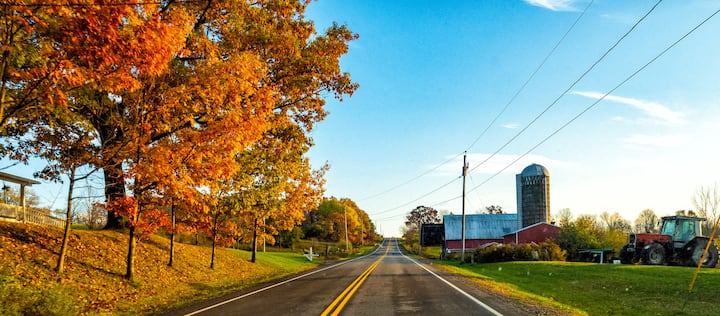 Country road in Western New York lined with autumn trees, passing farms and a red barn with silo under a blue sky