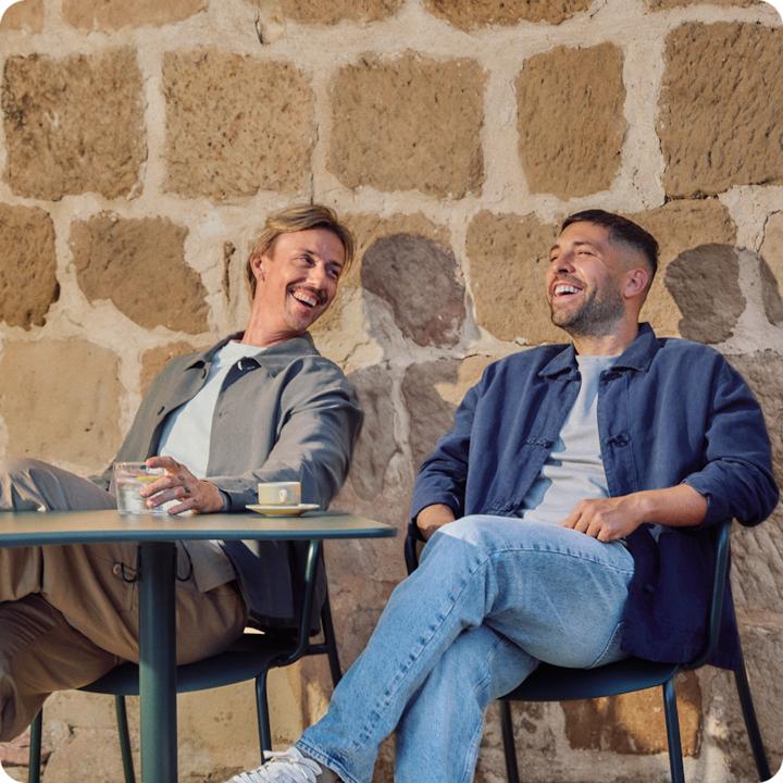 Footballers Jordi Alba and Guti (José María Gutiérrez) seated outside on a sunny terrace, sharing a laugh, with a rustic stone wall in the background.