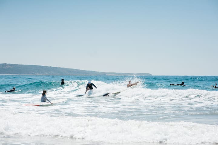 Unos surfistas surcan olas con crestas blancas en una playa australiana bajo un cielo azul despejado. La foto pone de manifiesto que las condiciones para practicar surf son seguras.