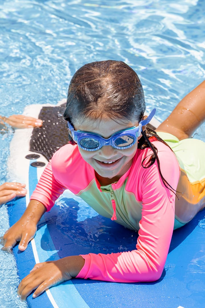 Un niña sonriente con gafas de bucear y un bañador de protección solar juega de forma segura sobre un flotador en una piscina de color azul claro.