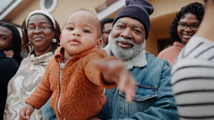 A smiling multigenerational family gathers outside a home as a baby reaches toward the camera.