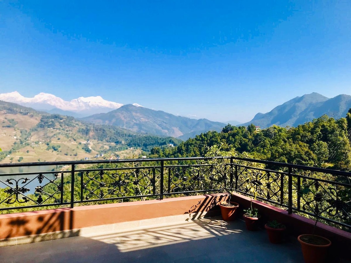 A spacious balcony is featured, showcasing a stunning view of the Himalayan Mountains and lush green hills. Potted plants line the railing, and the clear blue sky enhances the expansive vista, offering a serene environment for relaxation.