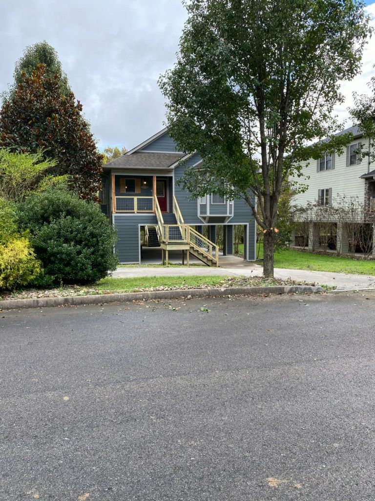 A two-story house is situated on a quiet street, featuring a gray exterior and a front balcony with wooden railings. Lush trees provide shade around the property, while a well-kept lawn enhances the welcoming appearance.