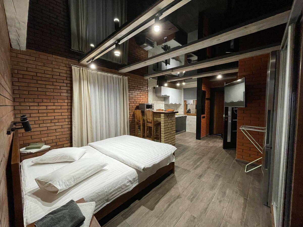 A spacious sleeping area is outfitted with a comfortable bed and crisp white linens. The warm tones of brick walls complement the modern kitchen and dining area in the background. Soft lighting is provided by overhead fixtures, creating a welcoming ambiance.