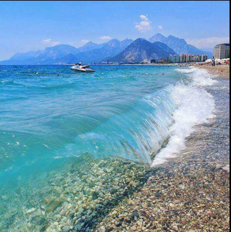 A serene beach landscape showcases gently rolling waves lapping against a pebbled shore. The clear blue water reflects the sky and reveals distant mountains, adding depth to the tranquil coastal scene.